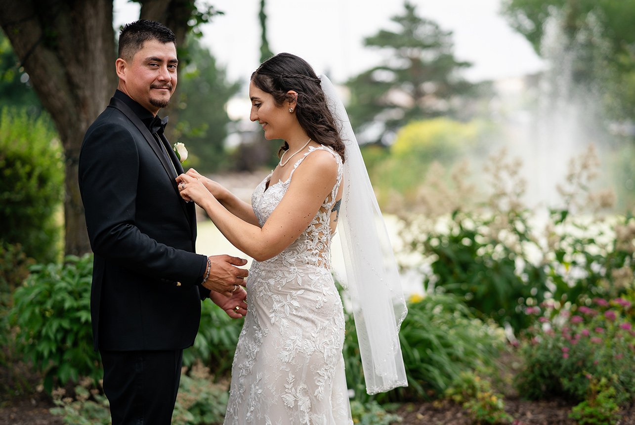 The groom looks over as the bride adjusts his vest and jacket suit, with lush foliage trees and a large water spring fountain in the backdrop at Sherwood Park's Greenland Botanic Gardens.