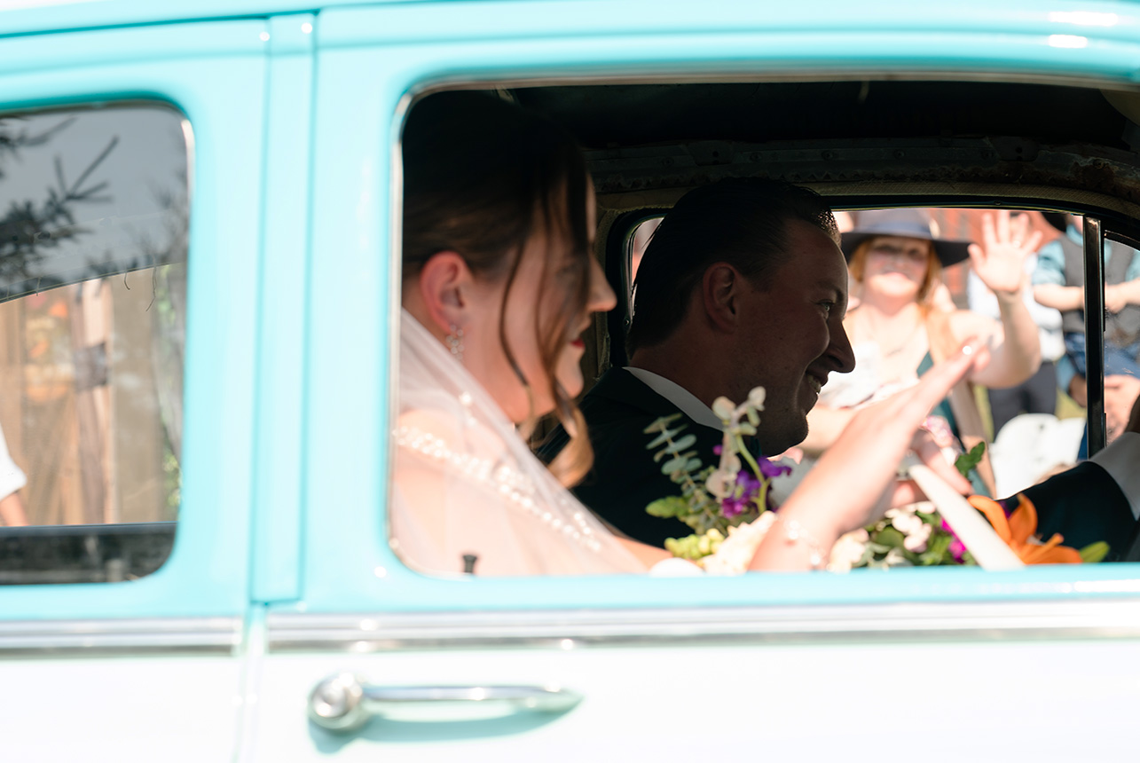 Groom drives his vintage car with the bride waving her bouquet, smiling at family as they drive off a rustic country road, a guest in a cowboy hat behind them
