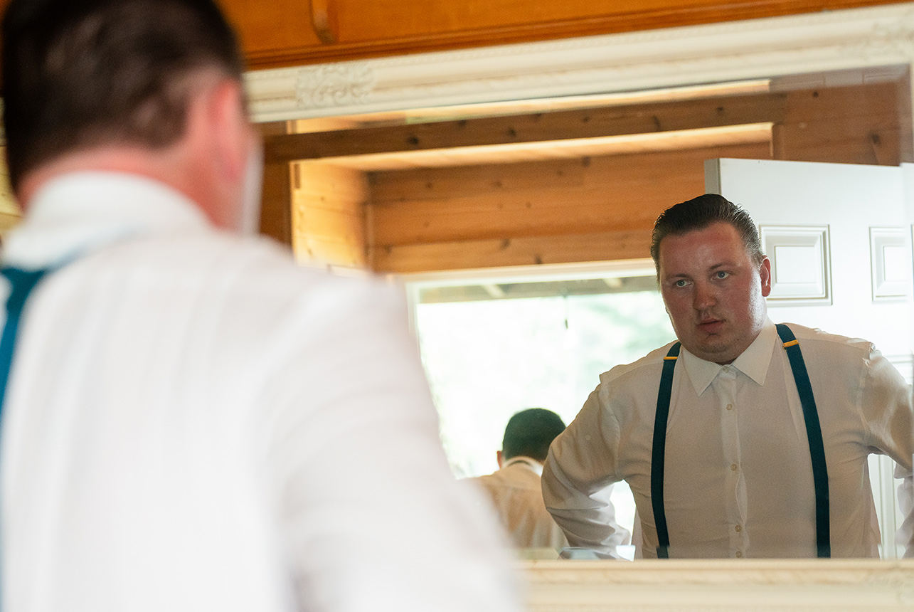 Groom gazes into the mirror, adjusting his suspenders as he prepares for his wedding day in the rustic cabin