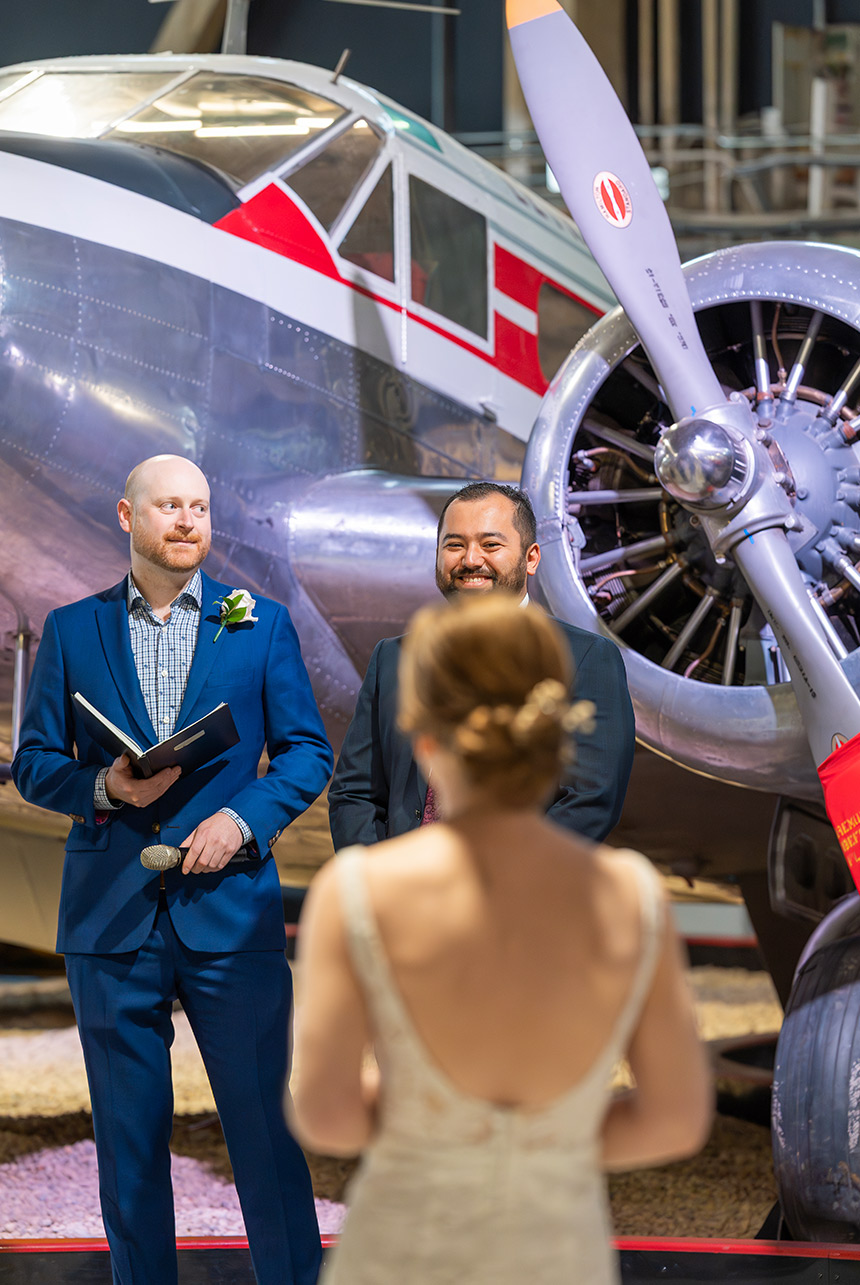 Groom at the front, in sharp focus, as the bride faces him in soft focus during an indoor ceremony with a vintage airplane backdrop