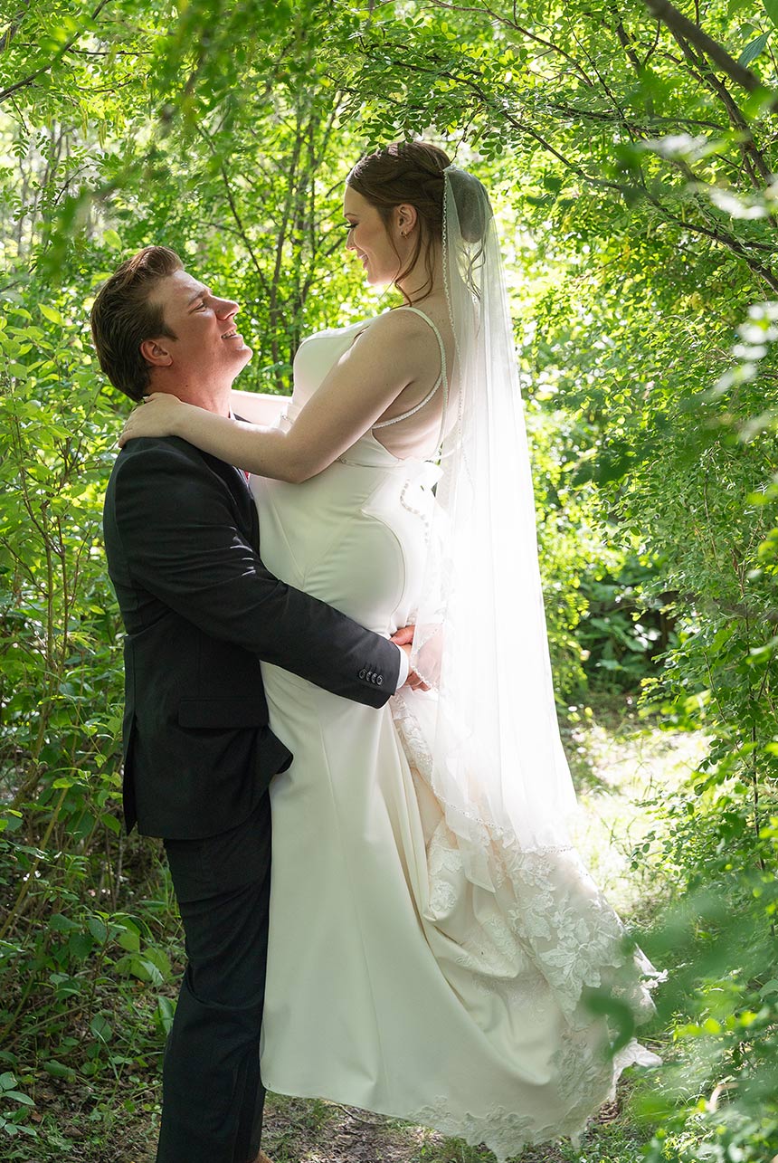 Groom lifting bride in forested trail at outdoor wedding