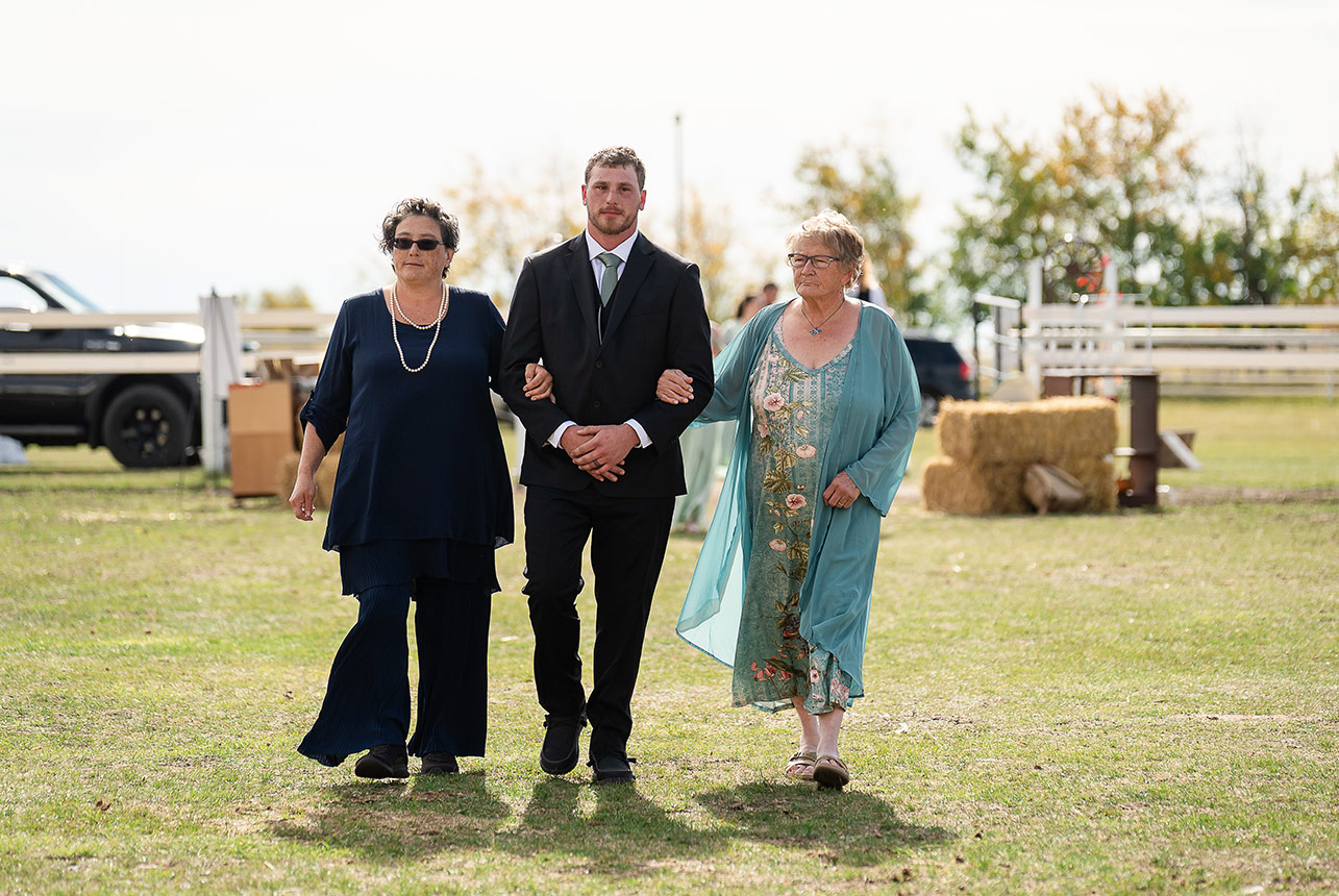 Groom escorted by his mother and grandmother at Finnegan Farms wedding, with blurred white fence and haybales in the background.