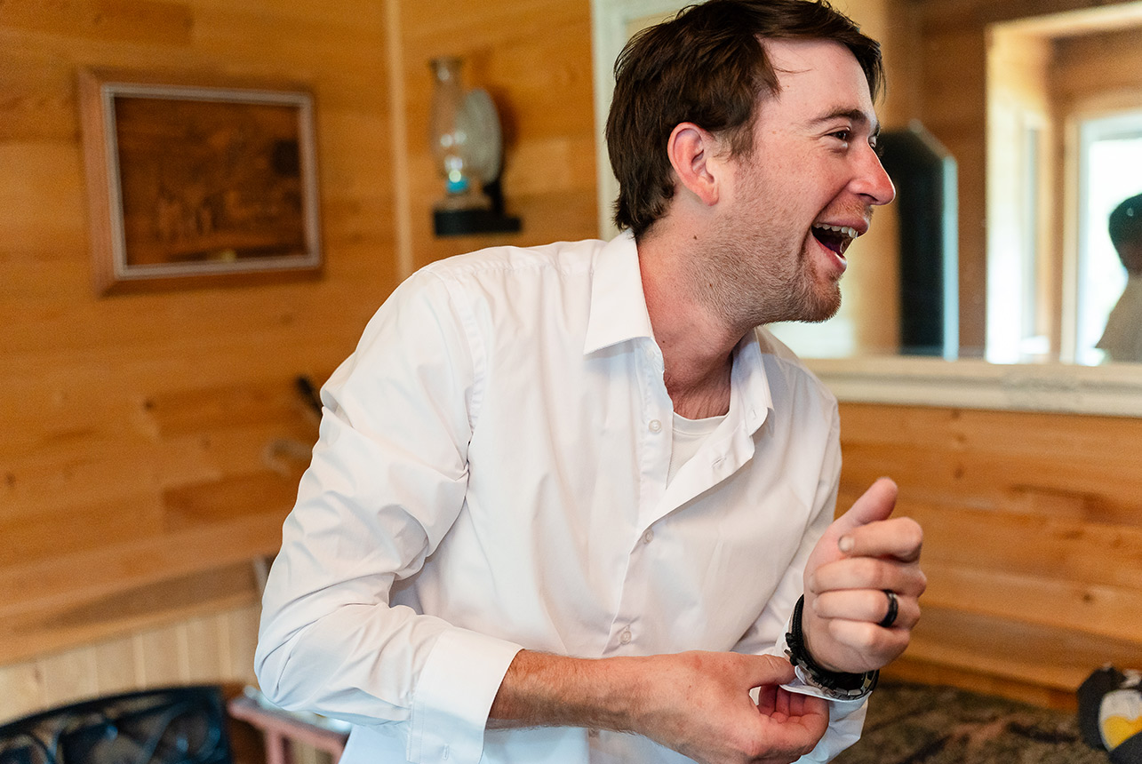 A groomsman bursts into laughter while buttoning his dress shirt in the rustic cabin on the morning of the wedding