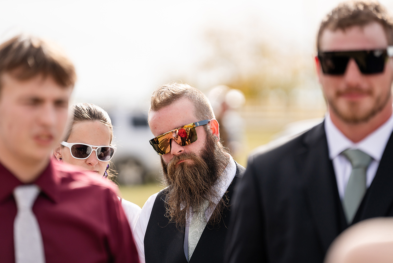 Close-up of a groomsman with a long beard and sunglasses reflecting the wedding scene
