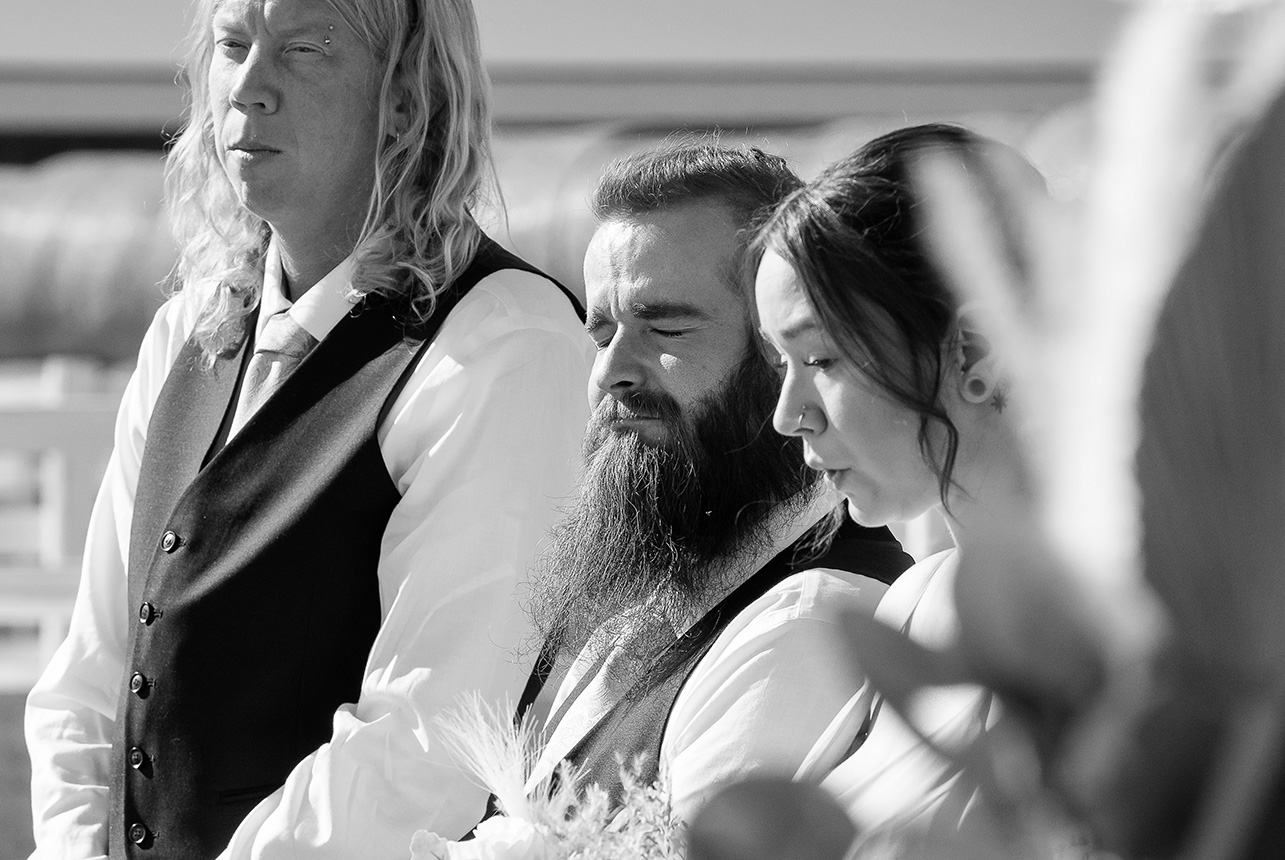 Two groomsmen and a bridesmaid react nervously and emotionally during the ceremony, with haybales and a barn roof in the background
