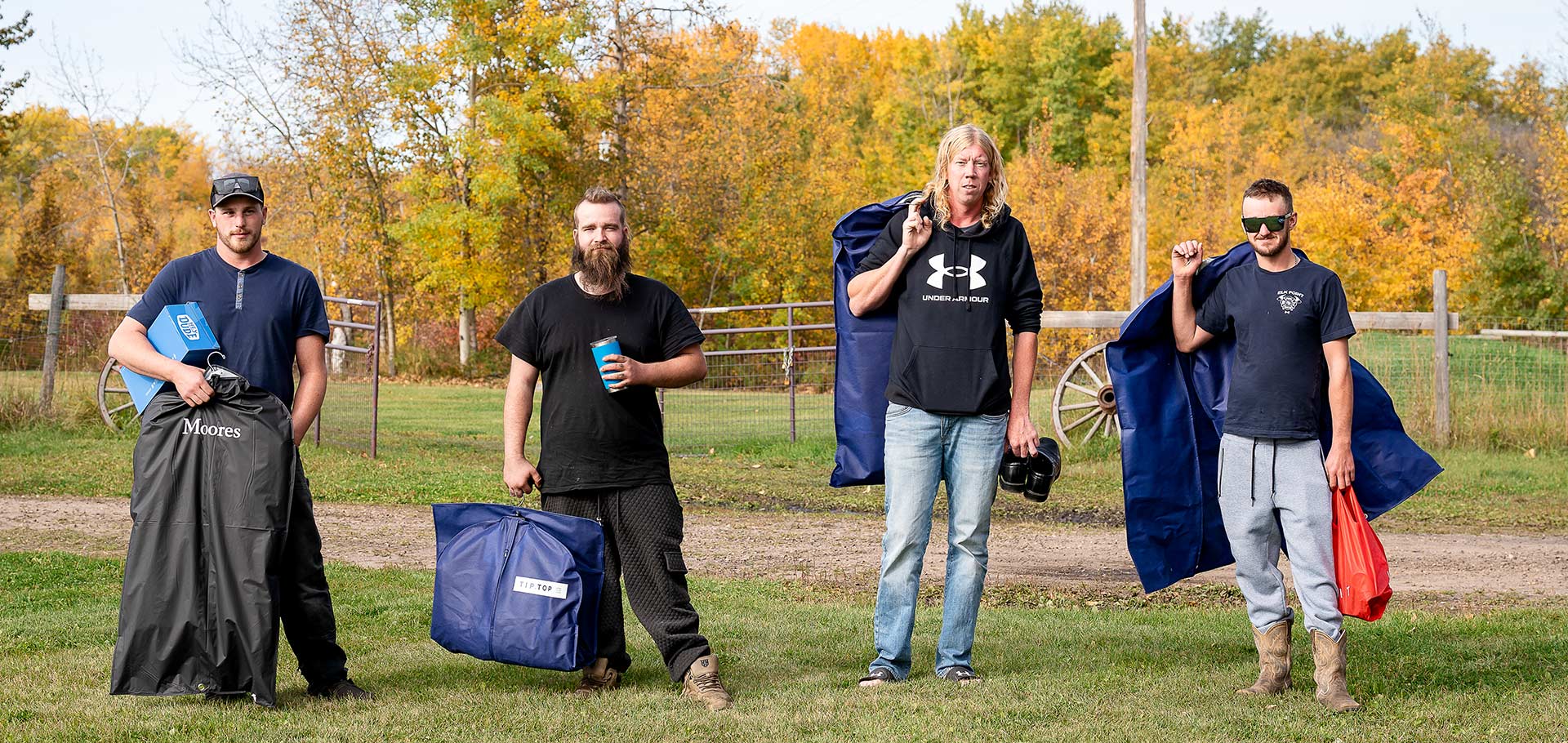 Four groomsmen walk along a gravel driveway, suits in bags over their shoulders, surrounded by autumn trees and a rustic wooden fence
