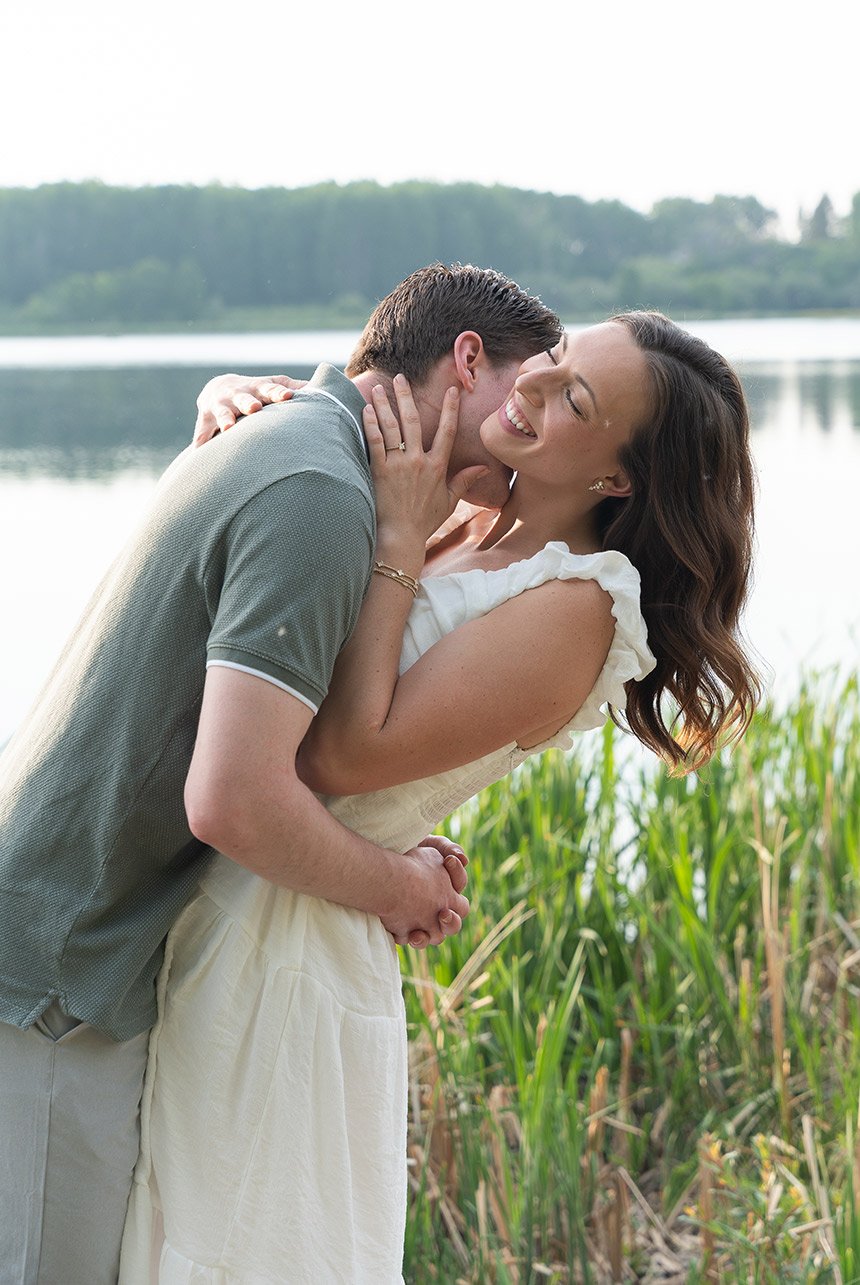 joyful-lakeside-kiss-during-engagement-session Bride-to-be laughs as her fiancé kisses her neck beside a peaceful lake