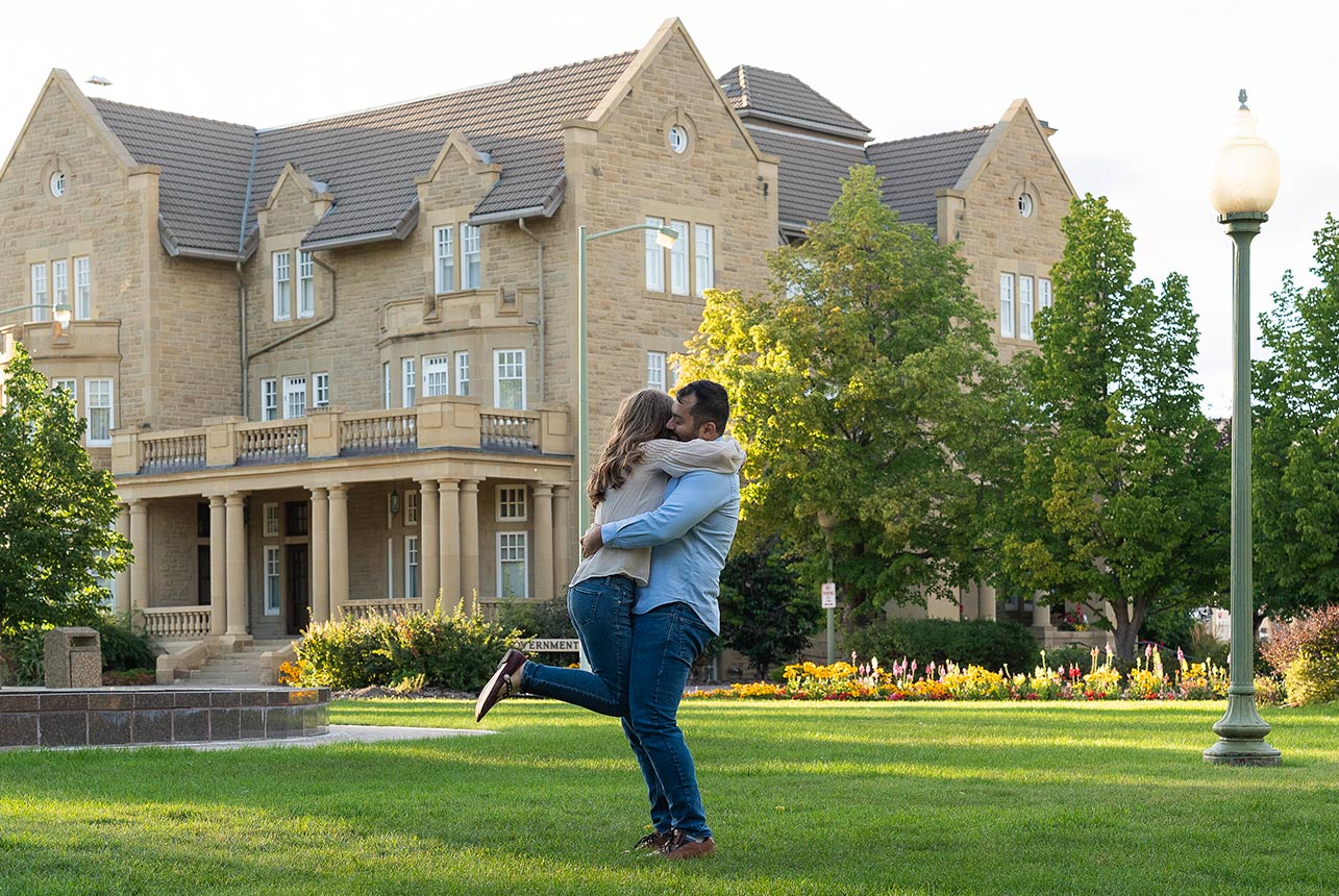 Couple hug on a sunlit lawn; she lifts one leg as they smile in front of a grand stone building