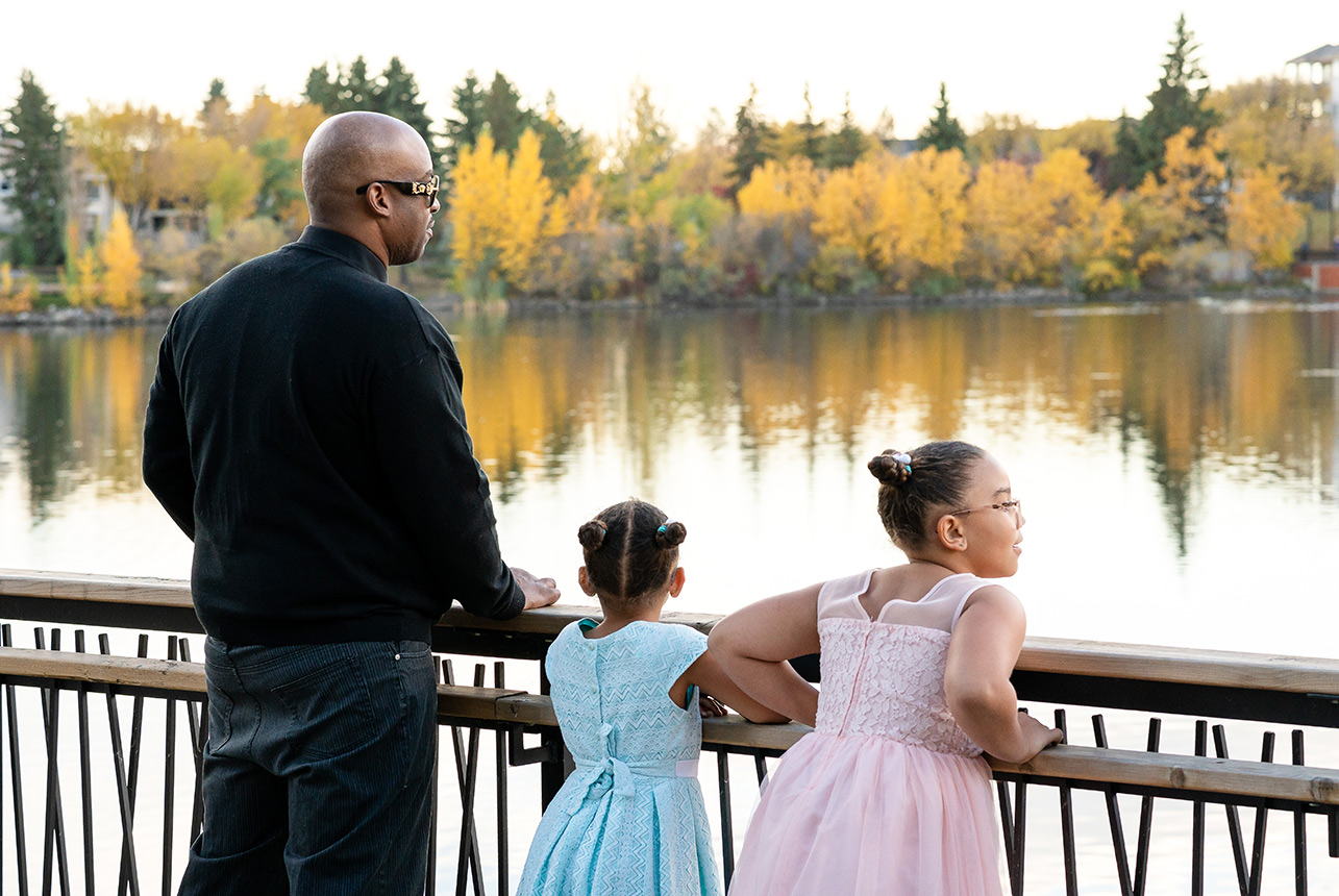 Father and two young daughters in pastel dresses admire autumn reflections at Beaumaris Park lake in Edmonton
