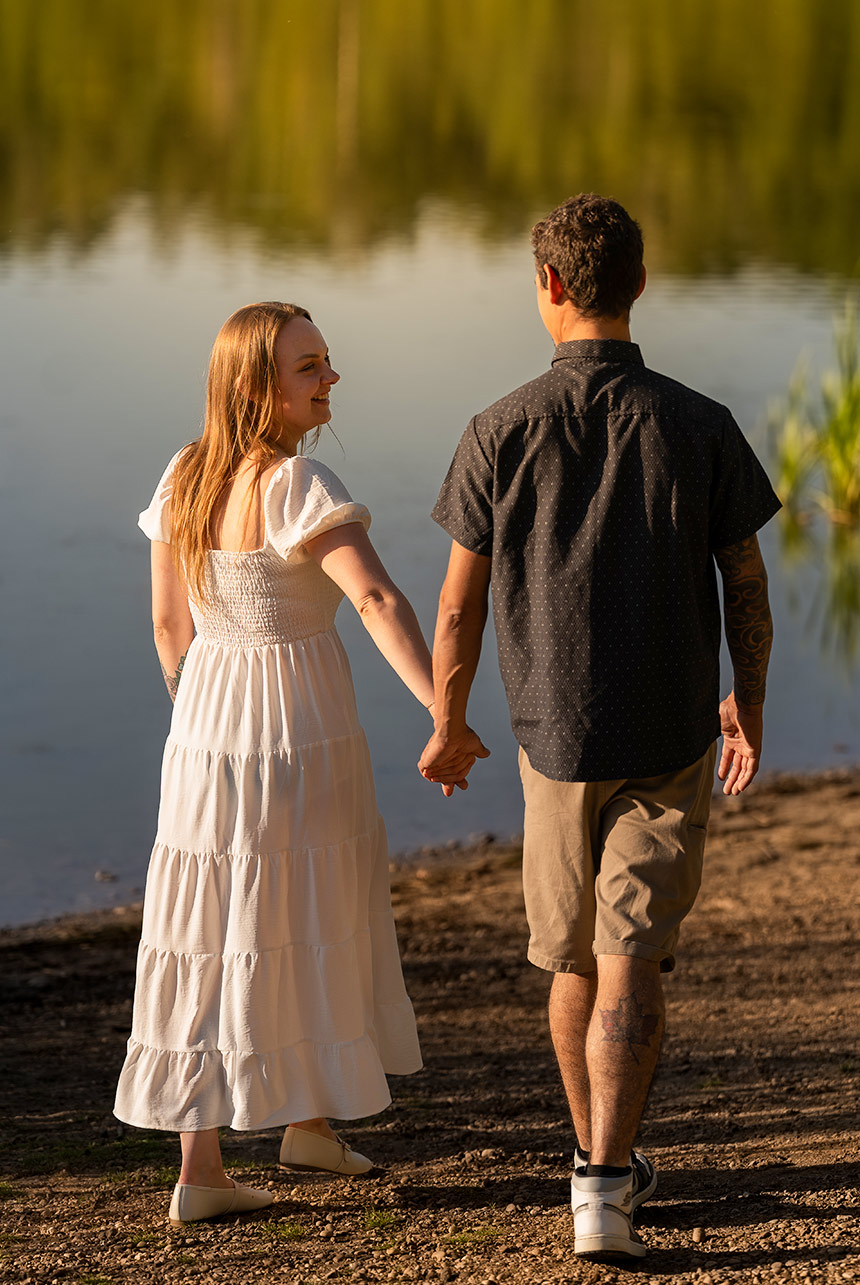 Couple walk hand in hand along the shore at Islet Lake; she looks back smiling in warm golden light