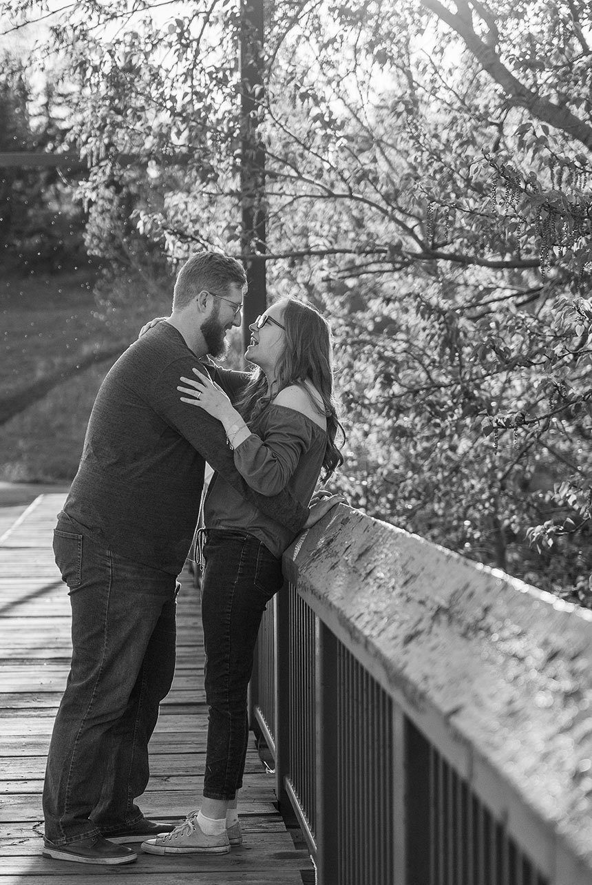 Couple laughing on a wooden bridge during golden hour with sunlight streaming through the trees at Rundle Park in Edmonton