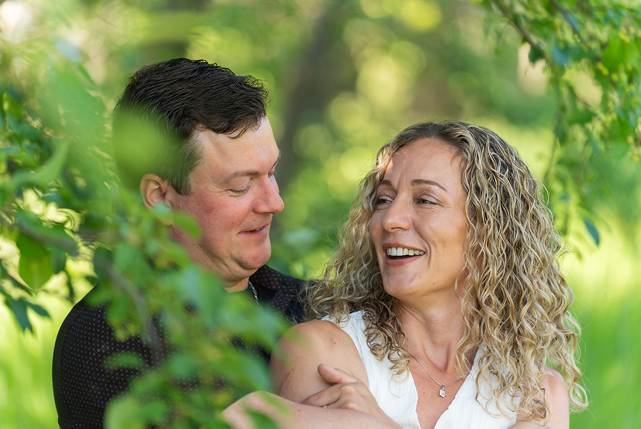 Couple laugh together in a close embrace, framed by soft green leaves and dappled light