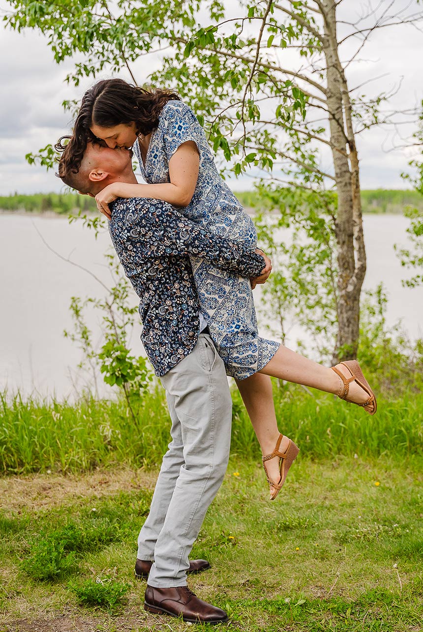 Man lifting bride-to-be in his arms for a passionate kiss with a lake and green forest backdrop in Elk Island, AB.