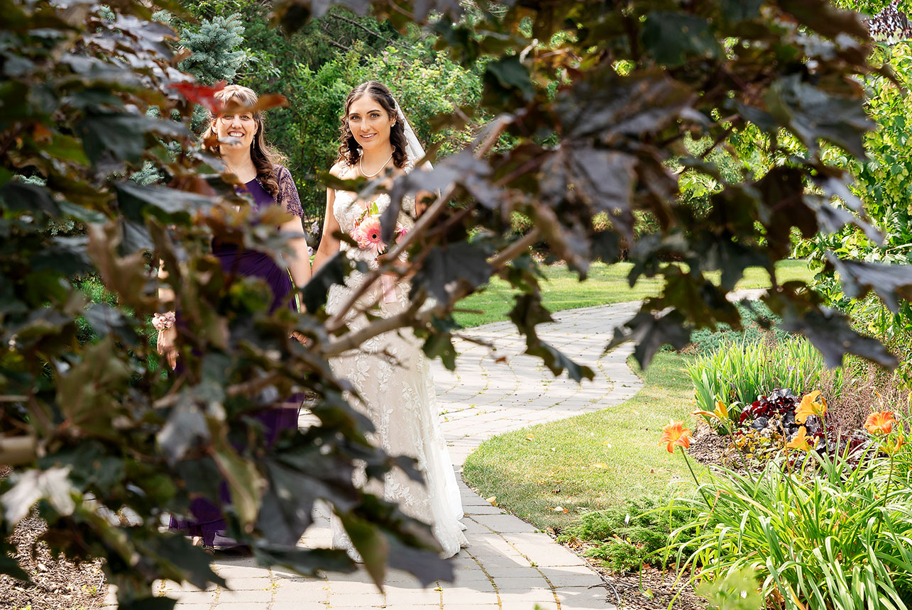A mother holding her bride's hand as they walk down the path towards the wedding aisles at Greenland Botanic Gardens, with tree leaves softly framing their faces.