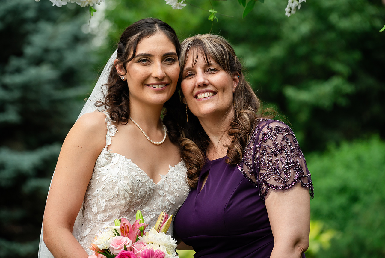 A mother giving a side hug to the bride holding her flower bouquet during the wedding ceremony at Greenland Botanic Gardens in Sherwood Park, near Edmonton.