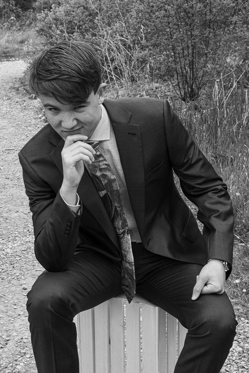 Young man sits on a wooden crate, dressed in his graduation suit and sporting a watch, with a contemplative expression on his face