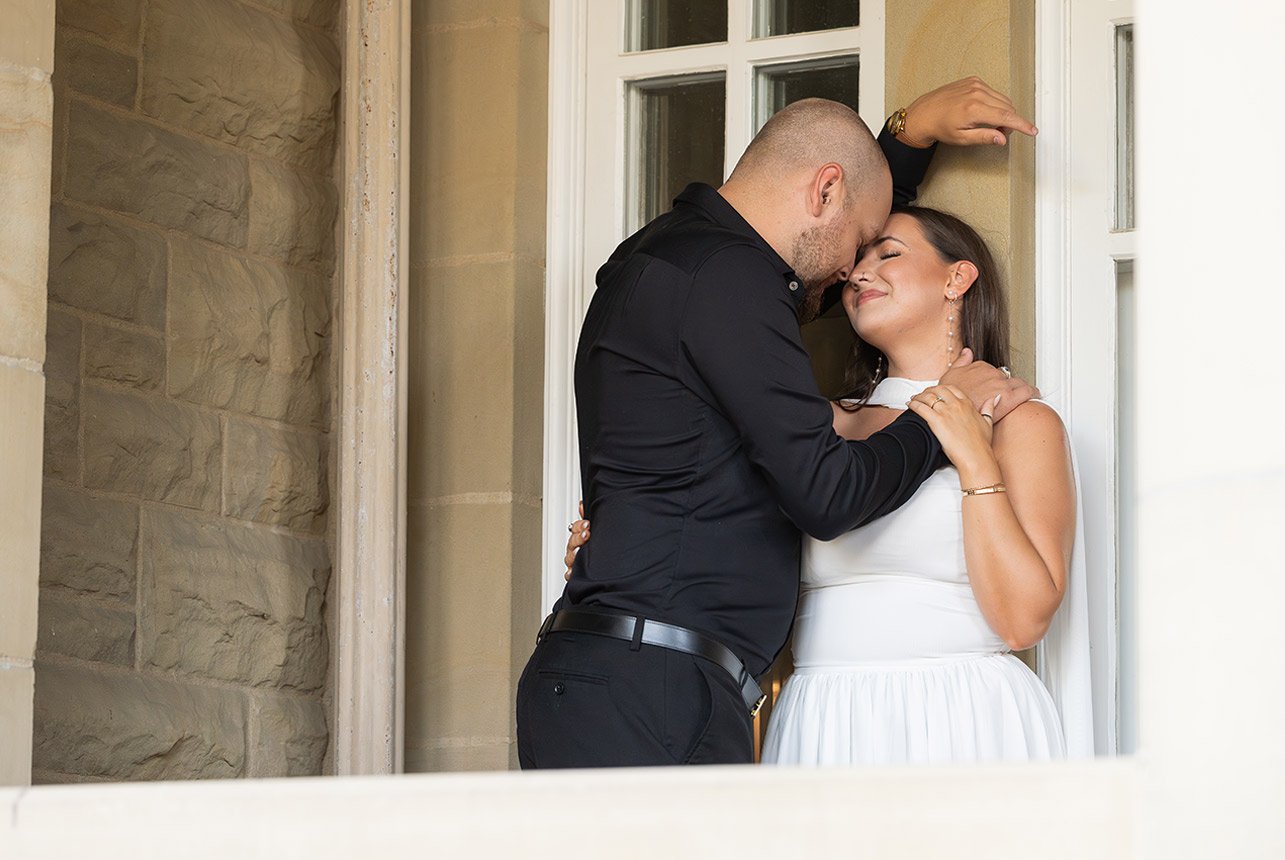 Couple nestles close in a stone alcove; his forehead rests against hers as she smiles with eyes closed, hand on his shoulder