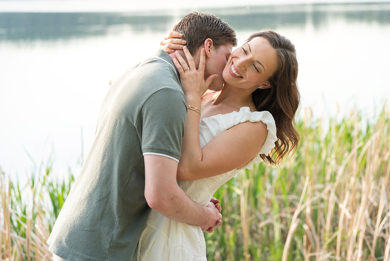 Woman smiling while partner kisses her cheek near a calm lake surrounded by reeds