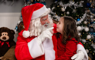 Santa points playfully as a young girl in a red sweater smiles on his lap beside the Christmas tree