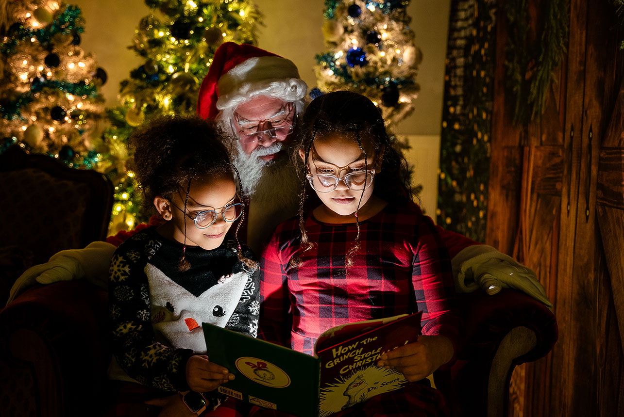 Two girls read a Christmas story as Santa leans in, their faces softly lit by the book