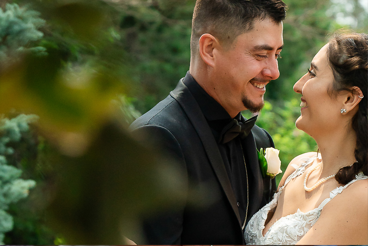 Side profile of a smiling couple gazing into each other's eyes, framed by blurred green tree leaves at Greenland Botanic Gardens in Sherwood Park, near Edmonton.