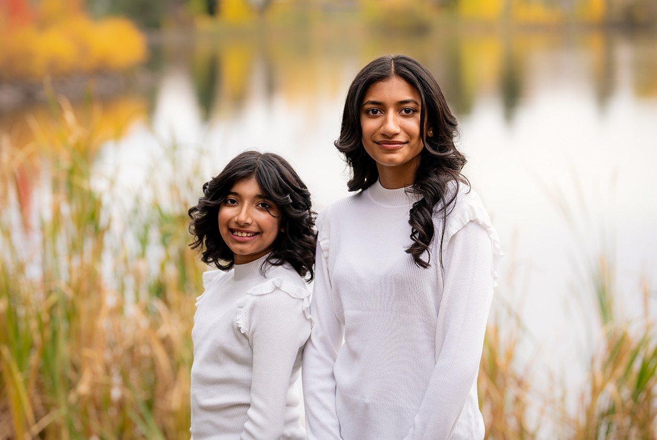 Two sisters standing in white sweaters by the lake, framed with soft golden fall tones