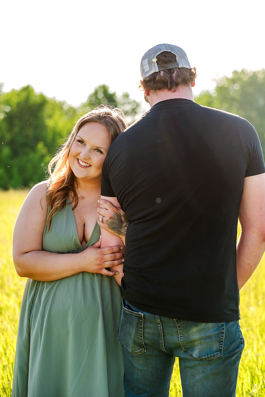 Woman smiling brightly while holding her partner’s arm in a golden sunlit field, engagement ring visible