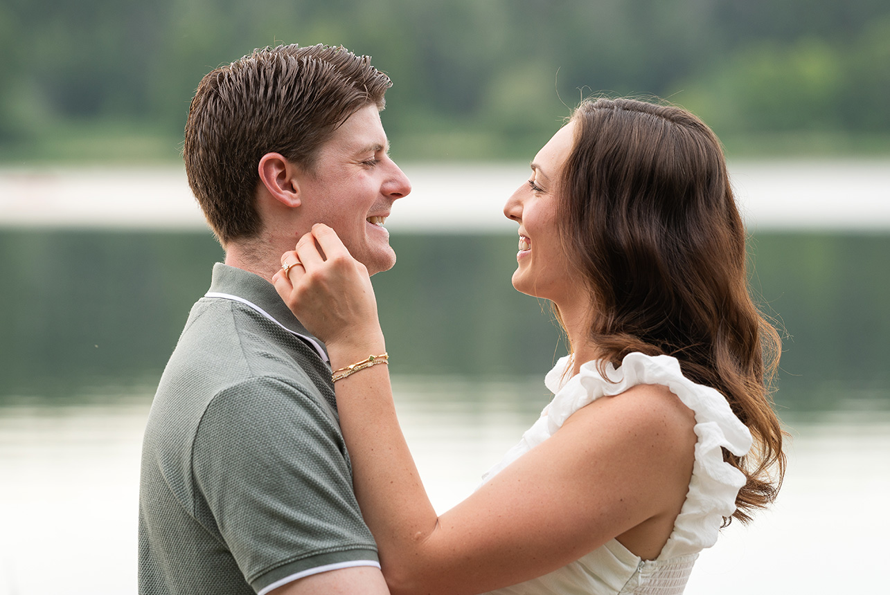 smiling couple face to face by still lake Couple faces each other, smiling as they share a quiet lakeside connection during their engagement shoot