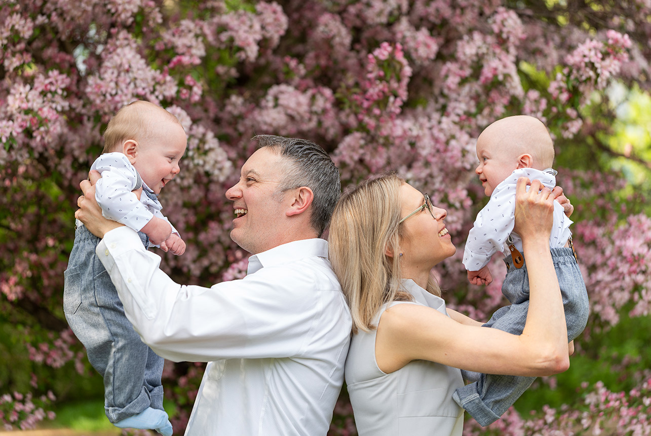 Parents lift twin baby boys in front of pink crabapple blossoms during a spring family session in Edmonton