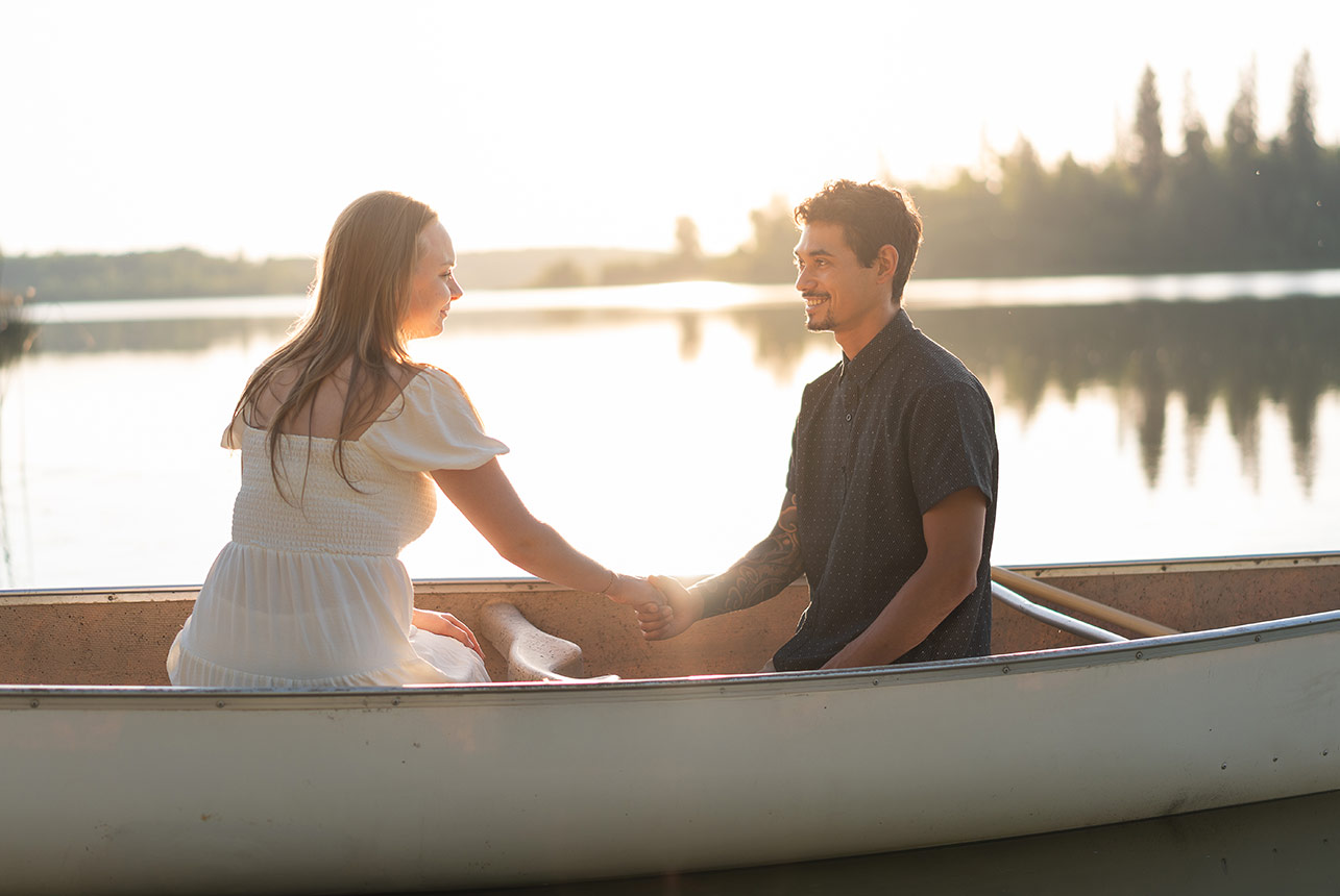 Couple sit facing each other in a canoe at sunset, holding hands with warm backlight and calm lake reflections