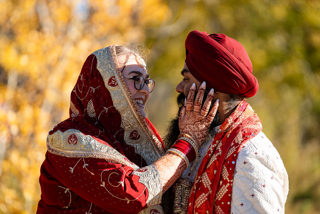 Bride in embroidered red and gold touches her groom’s face, henna-detailed hand, smiling in warm autumn light