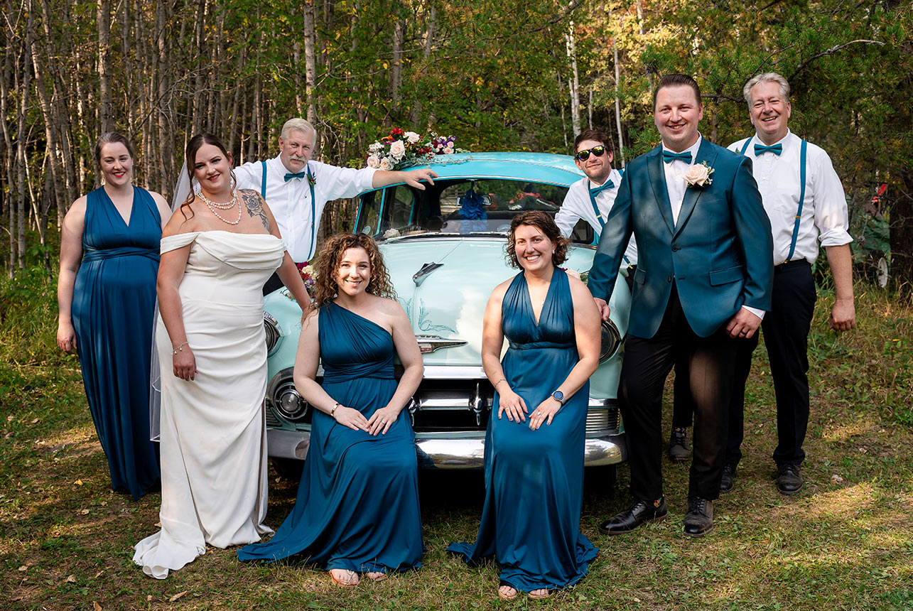 Color wedding photo of the wedding party surrounding a vintage car in a picturesque meadow, framed by a lush forest backdrop in Edmonton.
