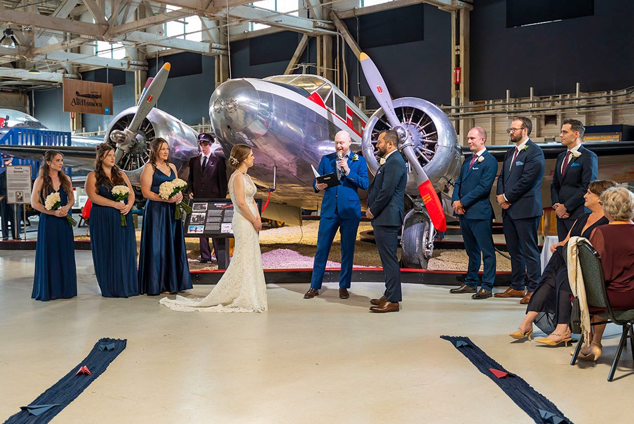 Bride and groom exchange vows at the Alberta Aviation Museum with a polished vintage aircraft behind them, wedding party lined up on both sides