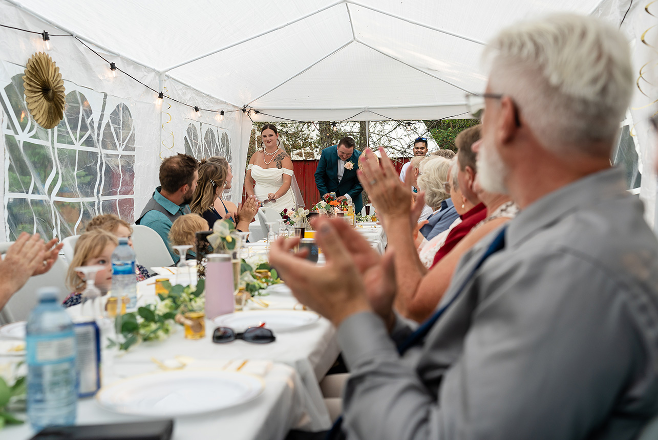 Wedding guests cheer and clap along a long, narrow reception table, welcoming the newlywed couple as they make their grand entrance