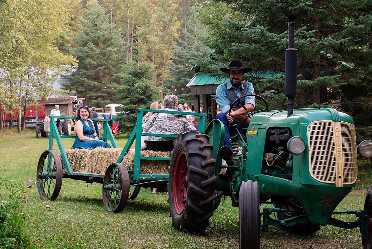 A cowboy drives a vintage steel-wheeled tractor, pulling a wagon of wedding guests enjoying a scenic hayride through the countryside