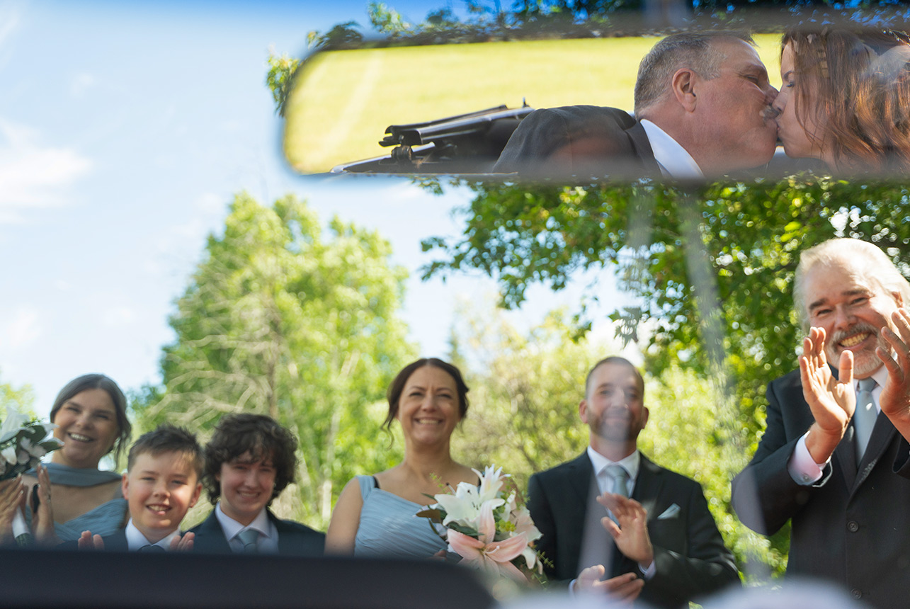 Bride and groom kissing in rearview mirror with wedding party cheering