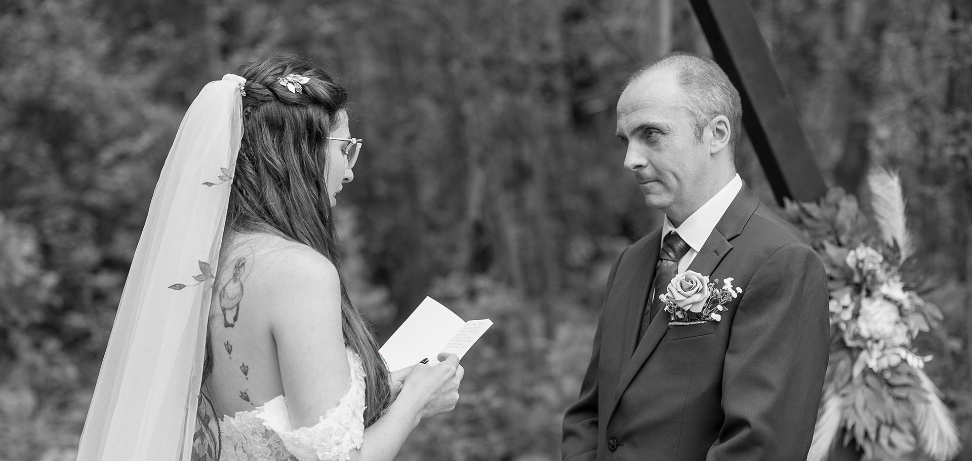 Bride reads handwritten vows to her groom during an intimate outdoor ceremony, black-and-white portrait framed by a simple floral arch