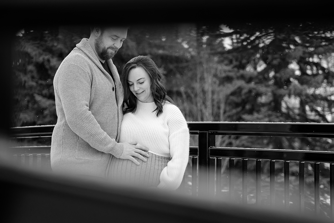 Expectant couple cuddles on a snowy bridge surrounded by evergreens during a winter maternity photoshoot in Edmonton