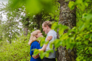 Woman in striking blue dress embraced by her future husband against a mossy tree, with a forest canopy at Emily Murphy Park.