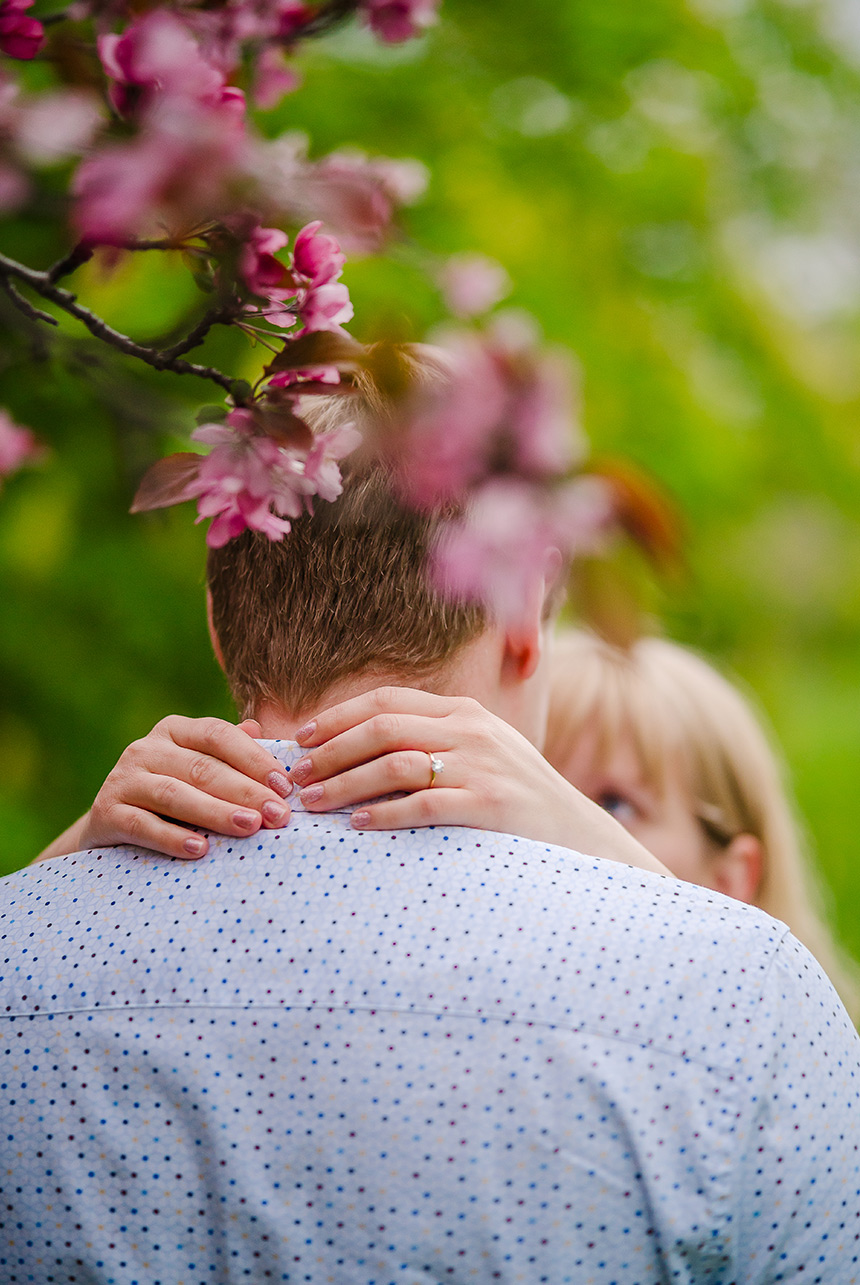 Woman wrapped around fiancé's neck, showing engagement ring under a blossoming Alberta Ornamental Crabapple Tree.